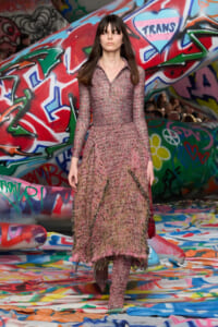 Model wearing a pink textured knit dress with fringe walking on a graffiti-covered runway as spectators watch.