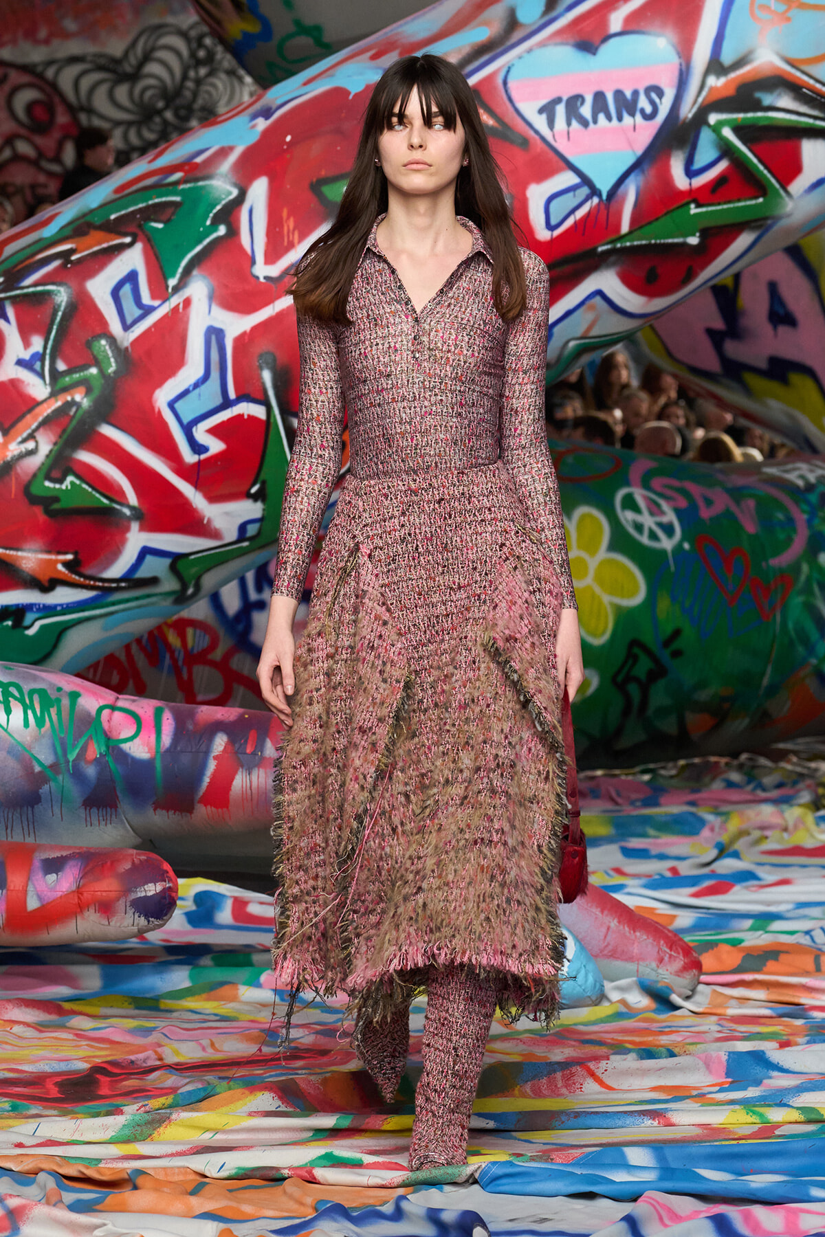 Model wearing a pink textured knit dress with fringe walking on a graffiti-covered runway as spectators watch.