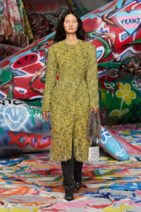 Runway model wearing a yellow textured coat, black pants, and pointed shoes, holding a gray fur bag and white clutch against a graffiti backdrop.