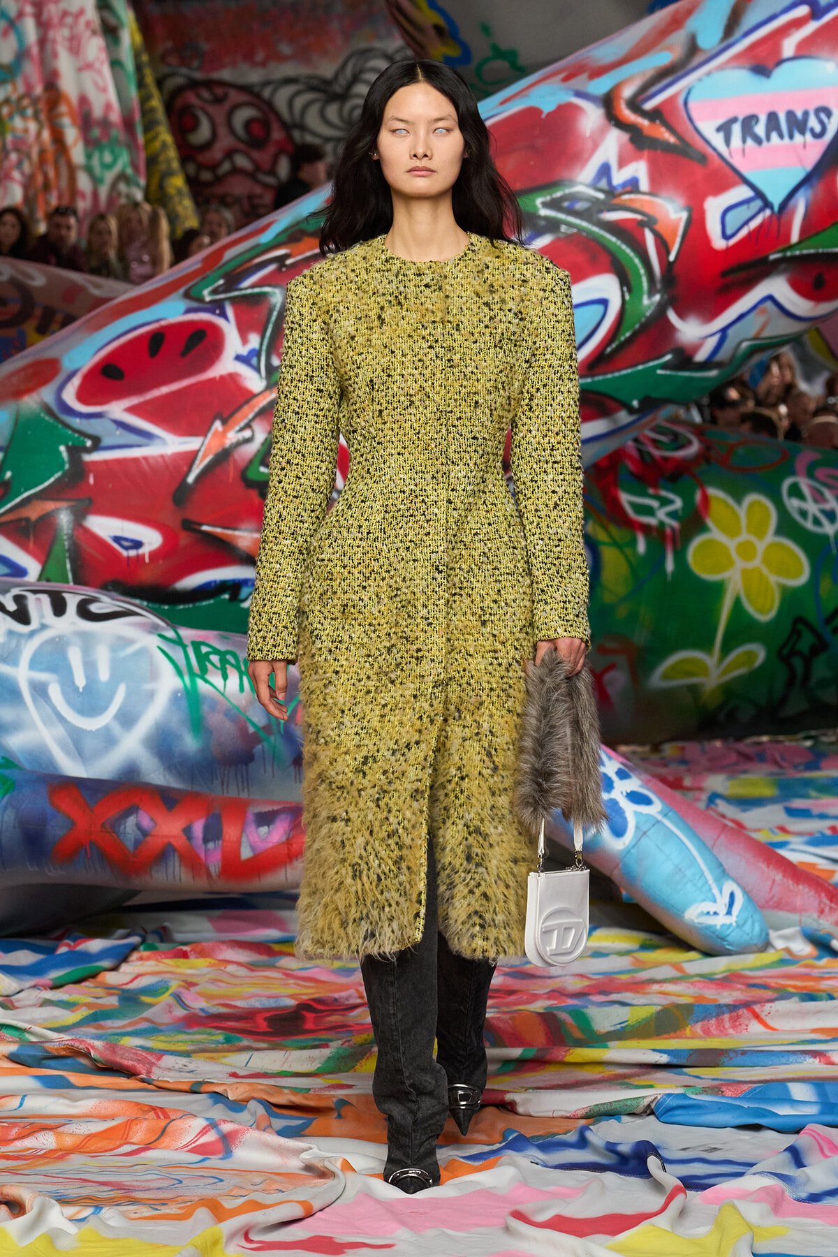 Runway model wearing a yellow textured coat, black pants, and pointed shoes, holding a gray fur bag and white clutch against a graffiti backdrop.