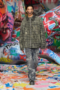 Model wearing a textured green jacket and patterned pants stands in a graffiti-covered tunnel.