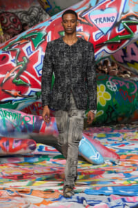 Male model walks a fashion show in a textured black jacket and gray cargo pants amid colorful graffiti structures in a street-art setting.