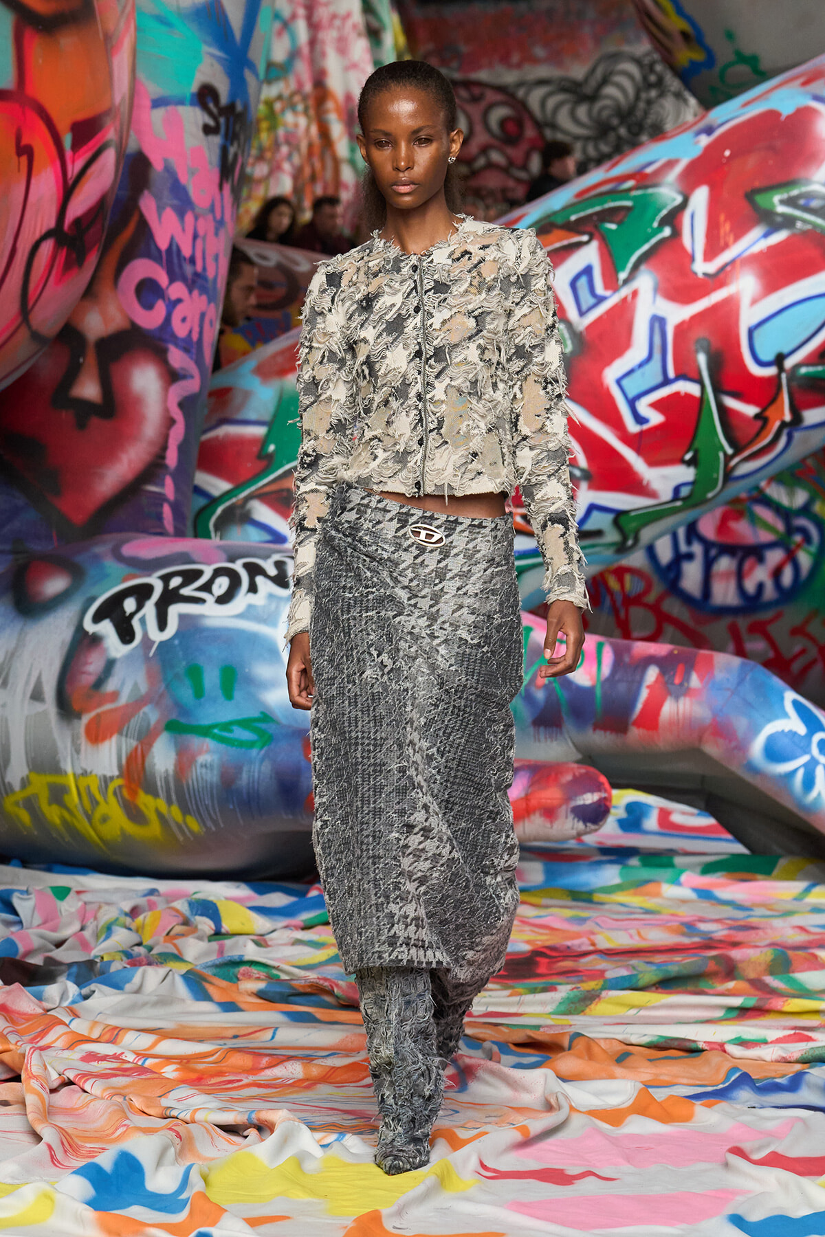 Model wearing a textured silver frayed jacket and matching metallic skirt with a belt, walking through a graffiti-covered tunnel.