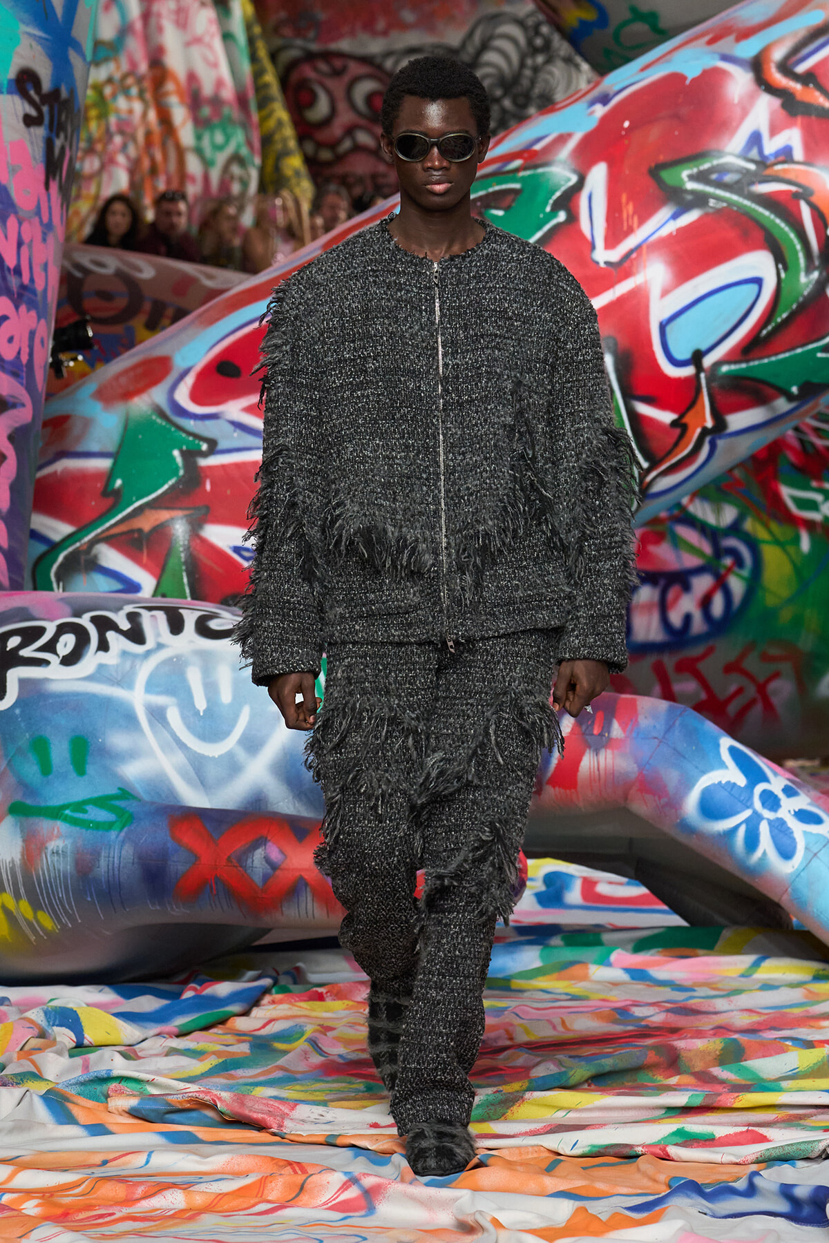 Model in a dark, fringed textured suit with sunglasses walks a graffiti-covered runway.