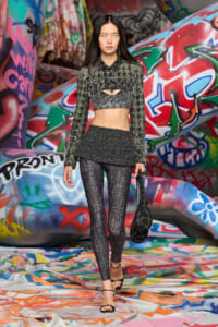 Model walking a runway in a metallic cropped top with long sleeves and matching glitter leggings, carrying a black textured bag, against a colorful graffiti backdrop.