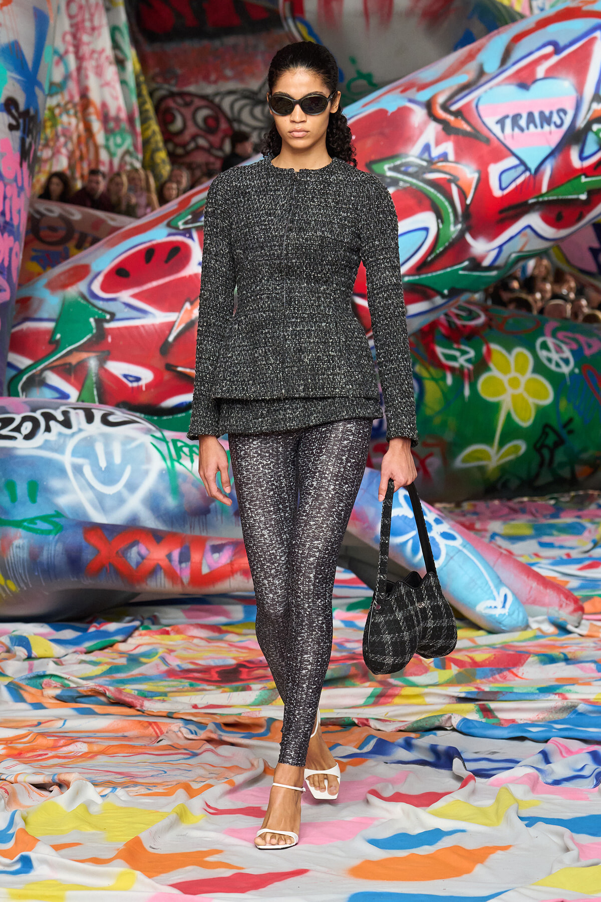 Model walks a runway in a metallic gray tweed set with matching leggings in a graffiti-filled venue. She wears sunglasses and white sandals, carrying a black plaid bag.