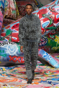 Male model wearing a grey-black textured zip-up jacket and matching pants walks on a colorful graffiti-covered runway backdrop.
