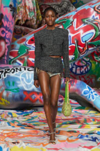 Runway model wearing a cropped gray tweed top and matching frayed shorts, carrying a bright lime-green beaded bag among graffiti-covered scenery.