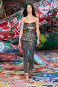 Model walks a grey strapless two‑piece outfit (peplum top with waist cutout and matching trousers) on a graffiti-covered runway with colorful background.