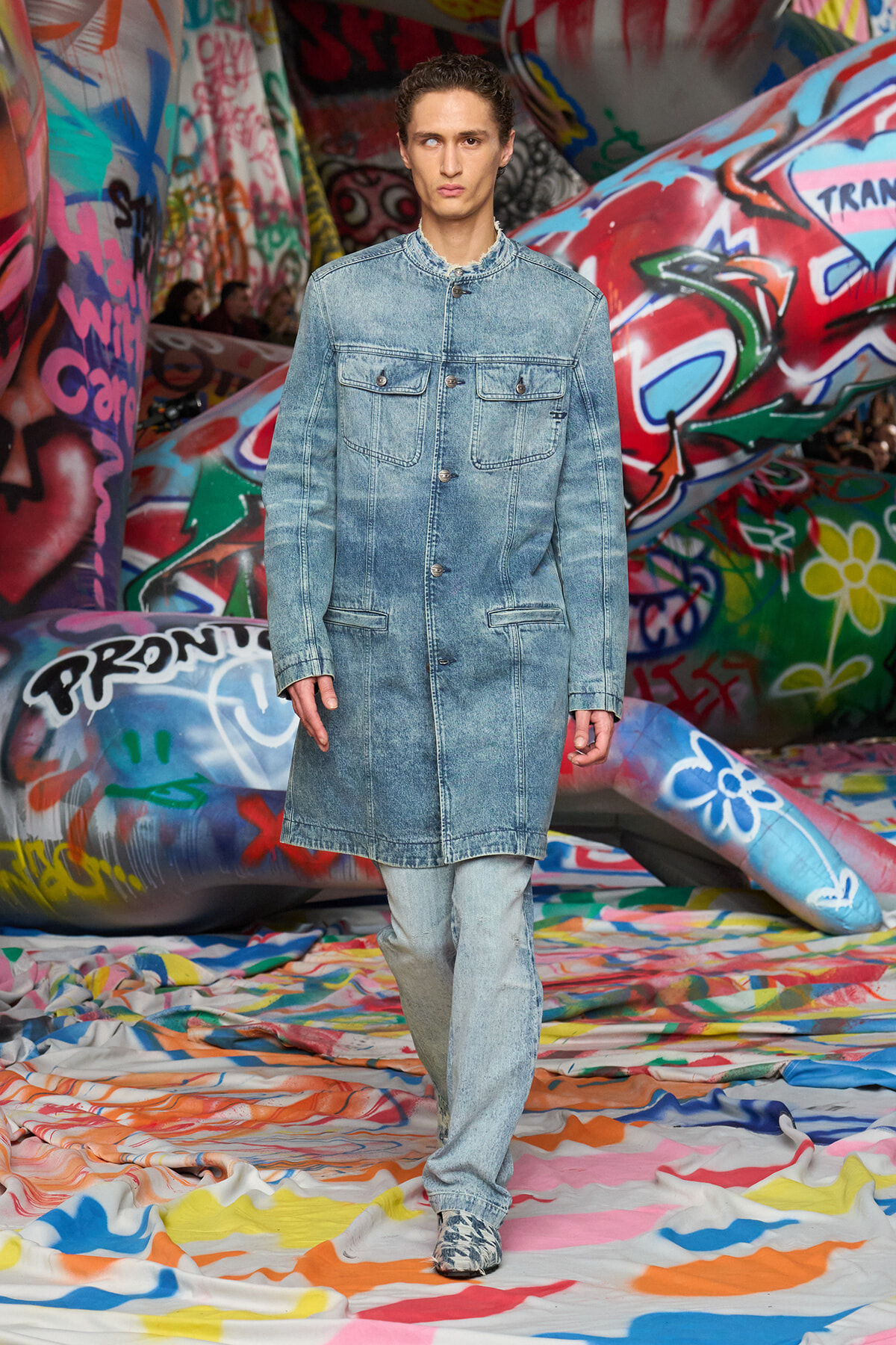 Male model walks a light denim coat over matching jeans down a colorful graffiti-covered runway, fashion show setting.
