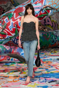 Female model walking in a graffiti-covered tunnel wearing a strapless charcoal corset top, light jeans, pink sandals, and carrying a black handbag.