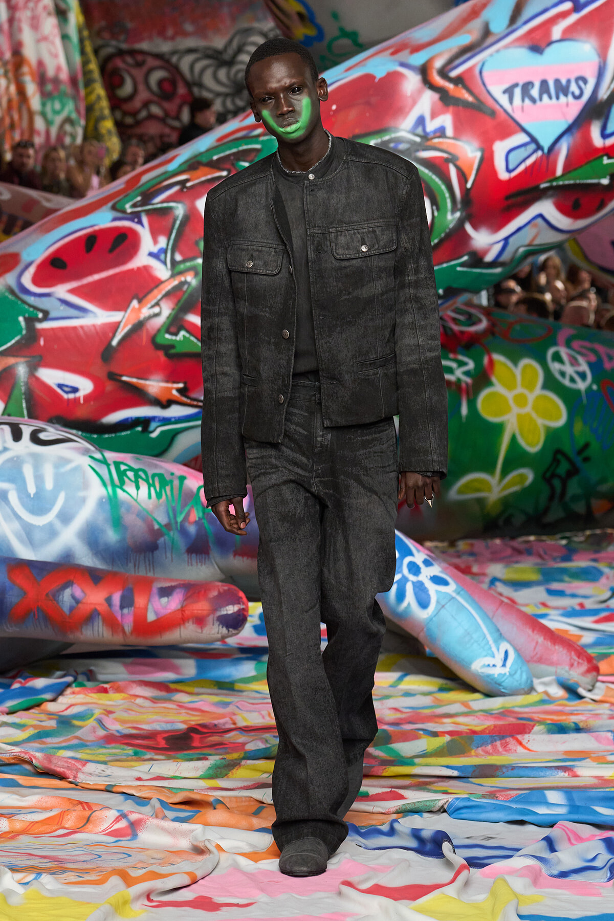 Male model walking a runway in a dark denim jacket and jeans with neon green facial paint, graffiti-covered backdrop behind him.
