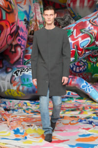 Male model wearing a knee-length gray coat over blue jeans walking through a graffiti-covered tunnel, looking straight ahead.