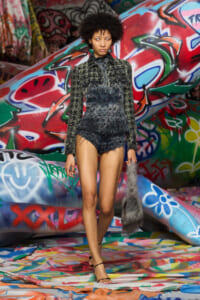 Model walks a colorful graffiti-covered runway in a dark, textured long-sleeve top and matching shorts, carrying a gray fur tail prop.