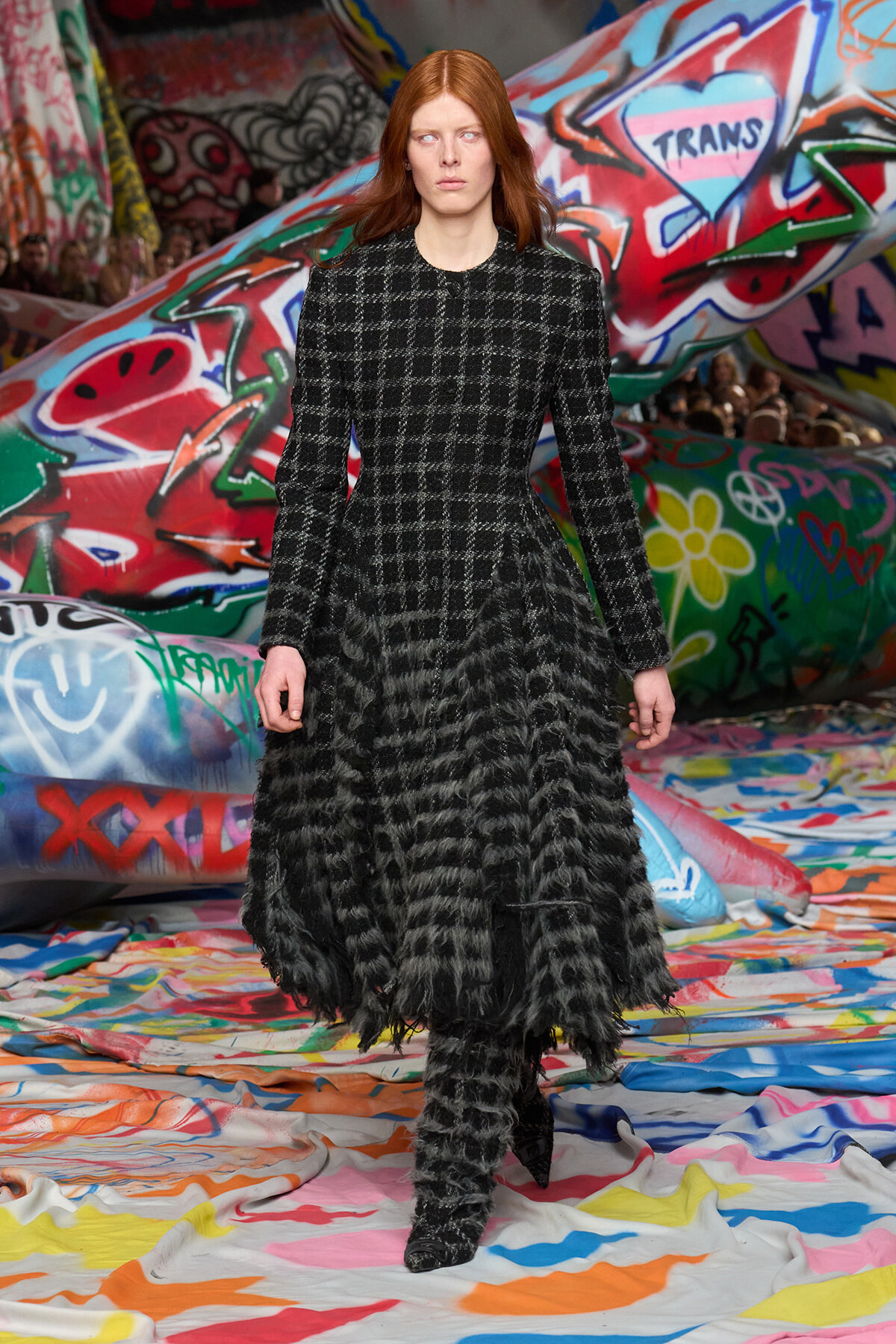 Model in a black-and-white checkered coat with a shaggy fur hem walks a graffiti-covered runway.