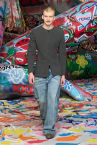Model walking a colorful graffiti-filled runway in a dark gray blazer and loose blue jeans.