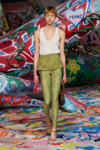 Model on a runway wearing a white sleeveless top with a lime-green, shimmering peplum and matching leggings, graffiti-covered backdrop behind her.