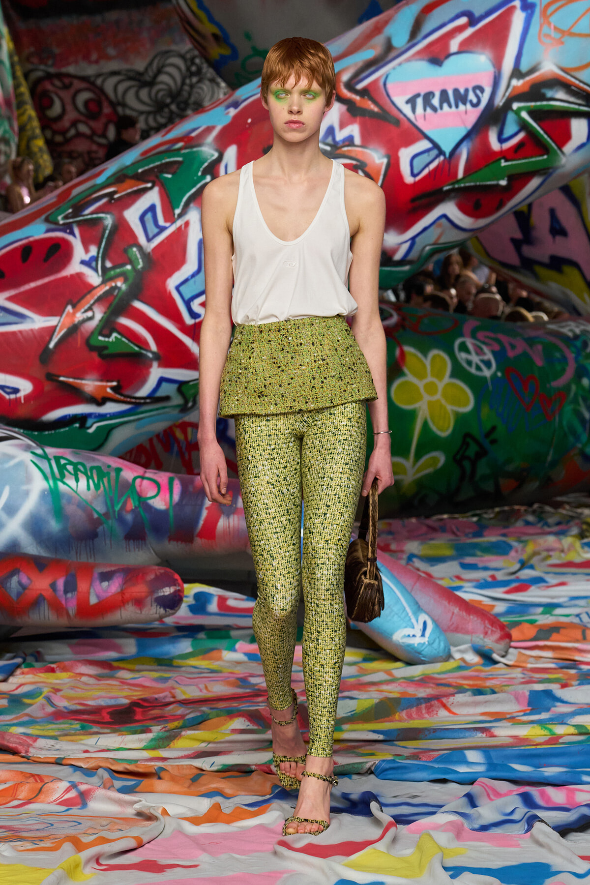 Model on a runway wearing a white sleeveless top with a lime-green, shimmering peplum and matching leggings, graffiti-covered backdrop behind her.
