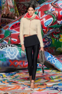 Model walking a runway in a cream cropped fleece jacket with red trim, black leggings, and a fur bag, against graffiti-covered backdrop.