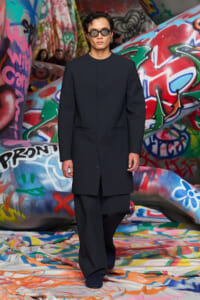 Male model in a black, minimalist overcoat and wide-leg trousers wearing dark sunglasses, walking on a graffiti-covered runway.