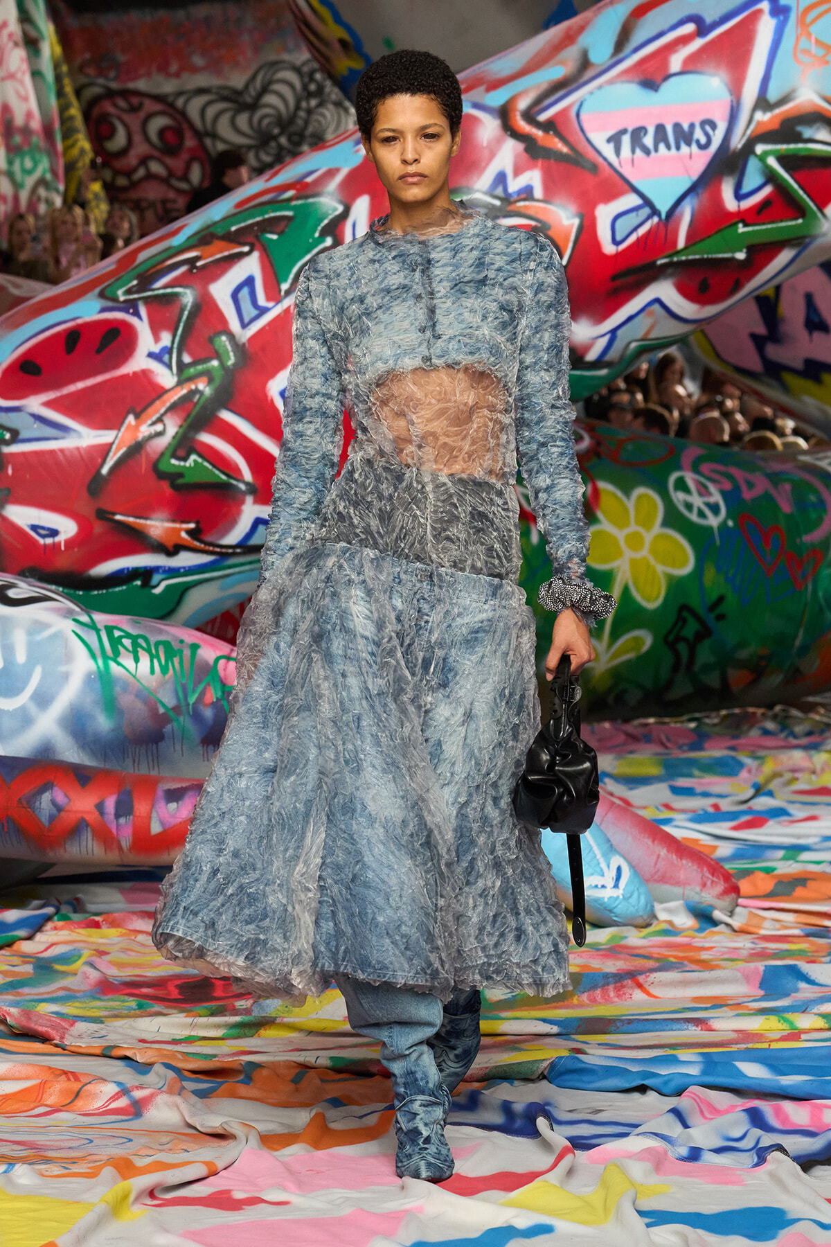 Runway model walking in a blue textured dress with a sheer midsection, blue boots, and a black bag, against colorful graffiti backdrop.
