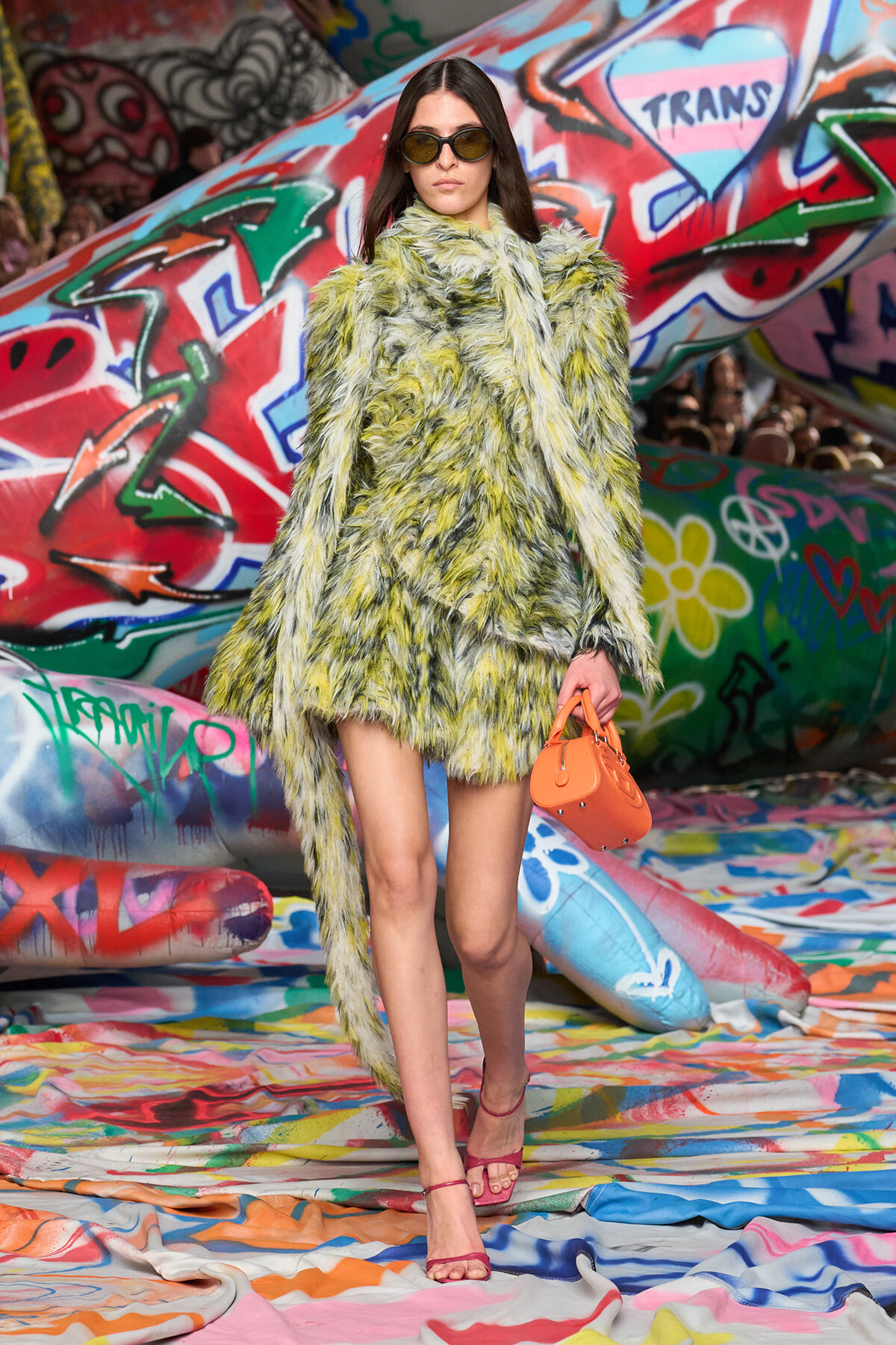 Model wearing a yellow-green faux fur coat and sunglasses, carrying an orange handbag against a graffiti-covered backdrop.