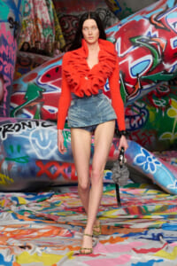 Model walking on a graffiti-filled runway in a bright red ruffled top and a denim mini skirt, striking pose.