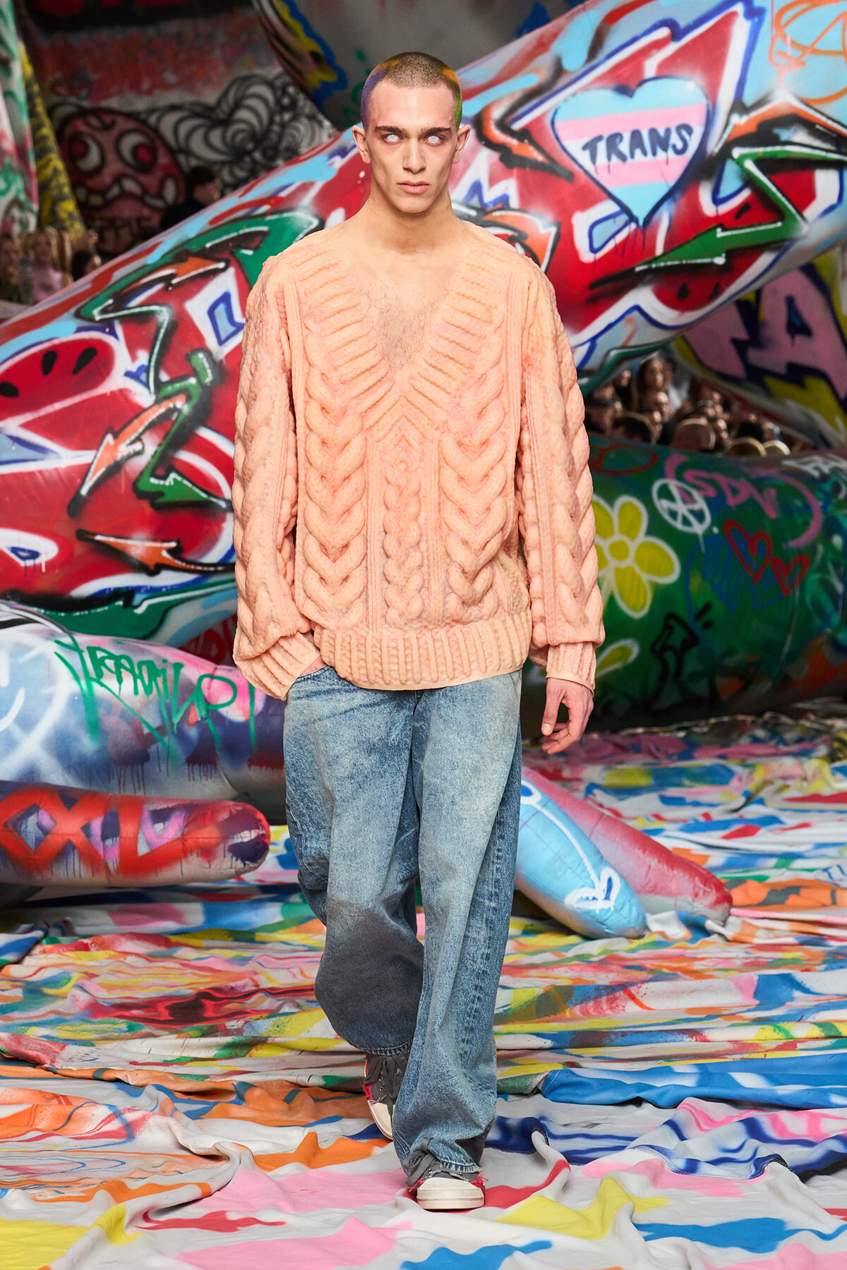 Male model walks down a graffiti-covered runway in a oversized peach knitted sweater and light-wash jeans.