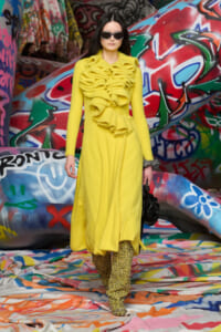 Model in a bright yellow ruffled coat and glittery pants stands before colorful graffiti walls, wearing sunglasses.
