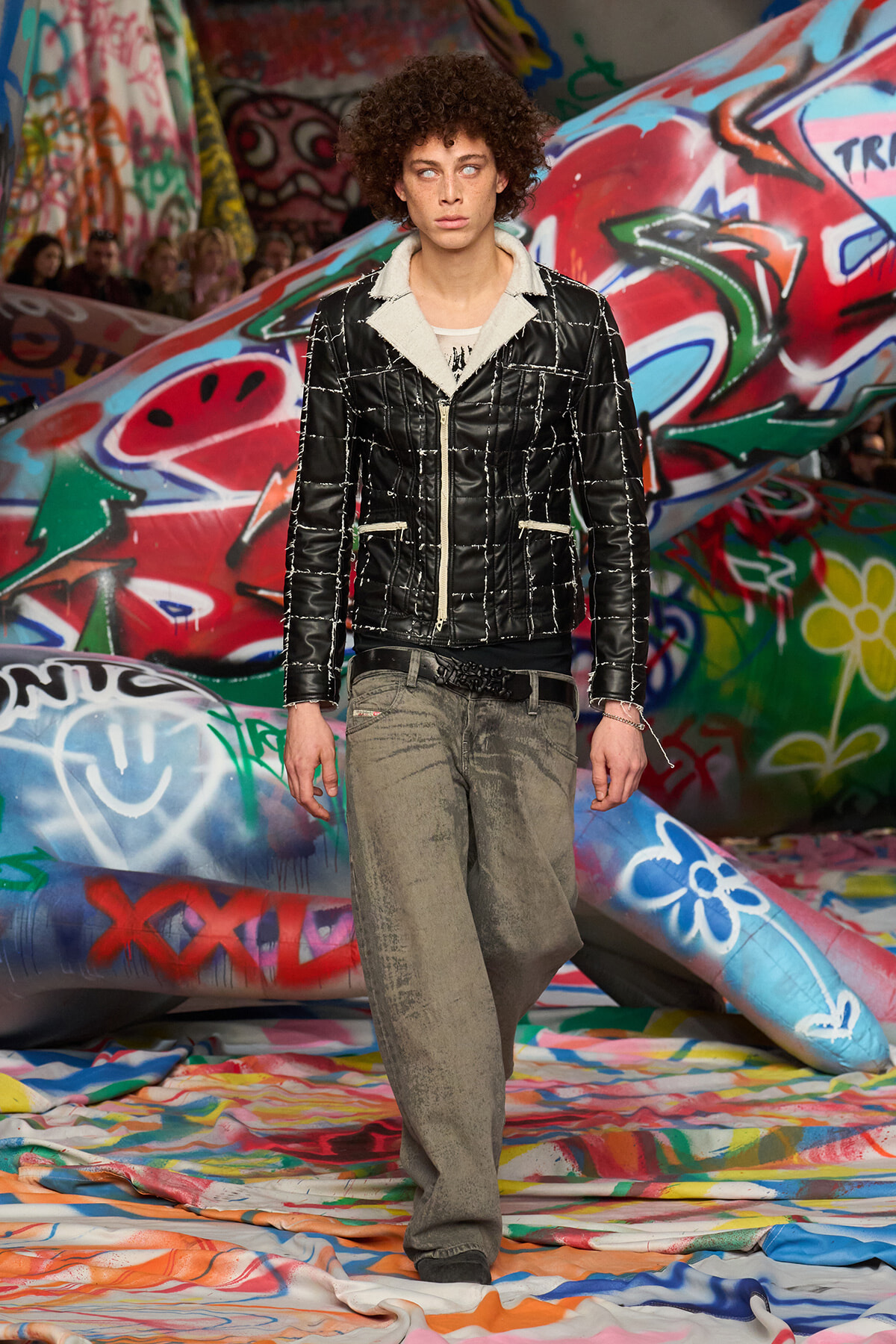 Model in a black quilted jacket with a light shearling collar and gray loose-fitting pants, walking through a graffiti-covered set.