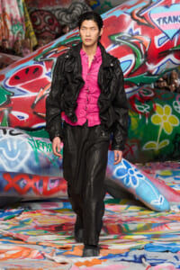 Male model in a pink button-down shirt under a black leather jacket walks a colorful graffiti-covered runway.