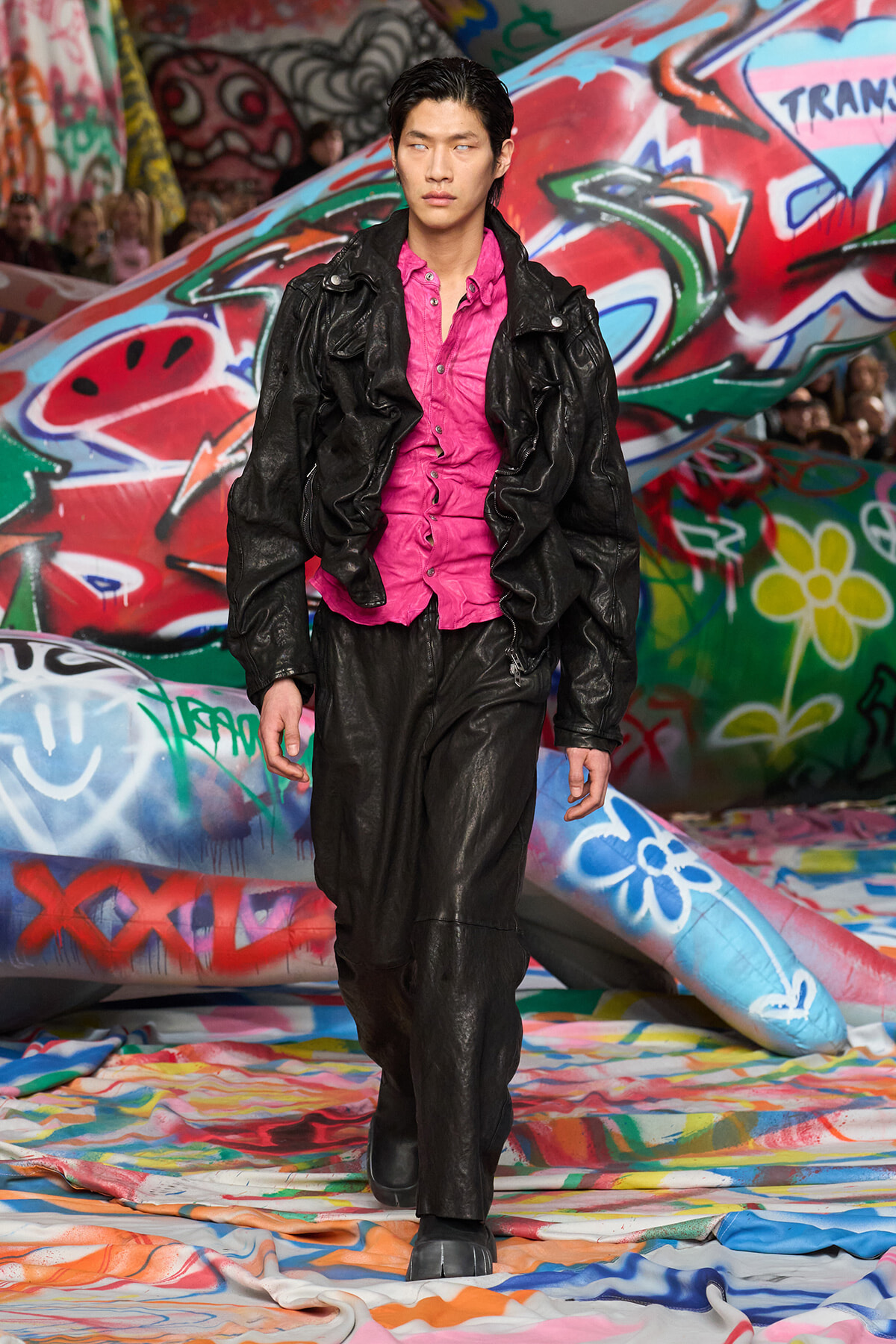 Male model in a pink button-down shirt under a black leather jacket walks a colorful graffiti-covered runway.