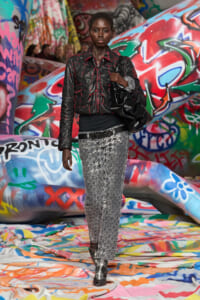 Model walking through a graffiti-filled tunnel in a black quilted jacket with red stitching, silver snakeskin pants, and a large black bag over the shoulder.