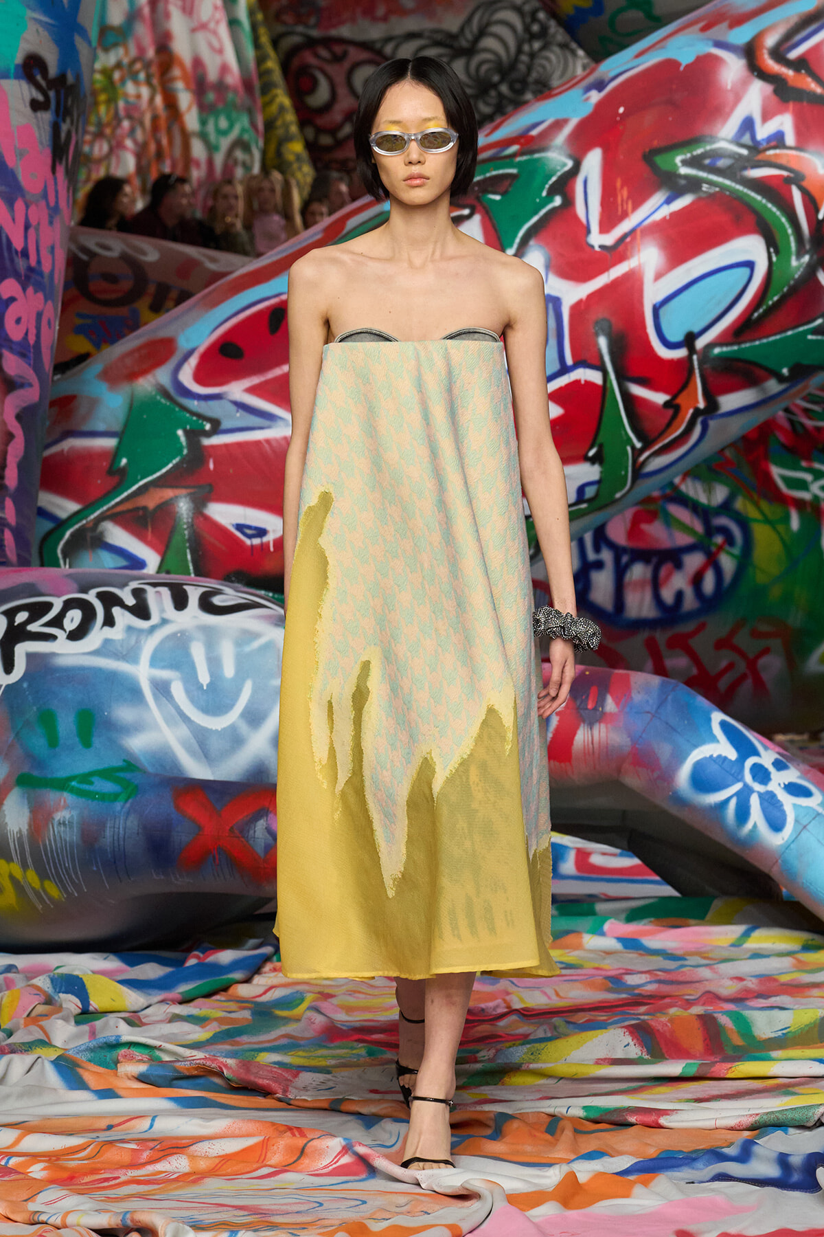 Model wearing a strapless pastel dress with a pale chevron pattern, standing against a graffiti-covered backdrop.