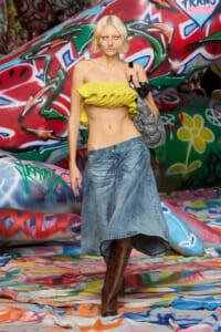 Model posing in a bright yellow ruffled crop top, a denim midi skirt, and brown boots in front of colorful graffiti art on a runway.