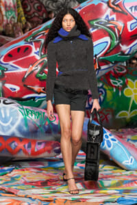 Model walking a graffiti-covered runway in a gray knit sweater with blue collar, black shorts, sandals, and a black bag at her side.