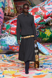 Model on a graffiti-covered runway wearing a dark zip-front jacket, red sunglasses, a wide gold belt, navy trousers, and a black bag.