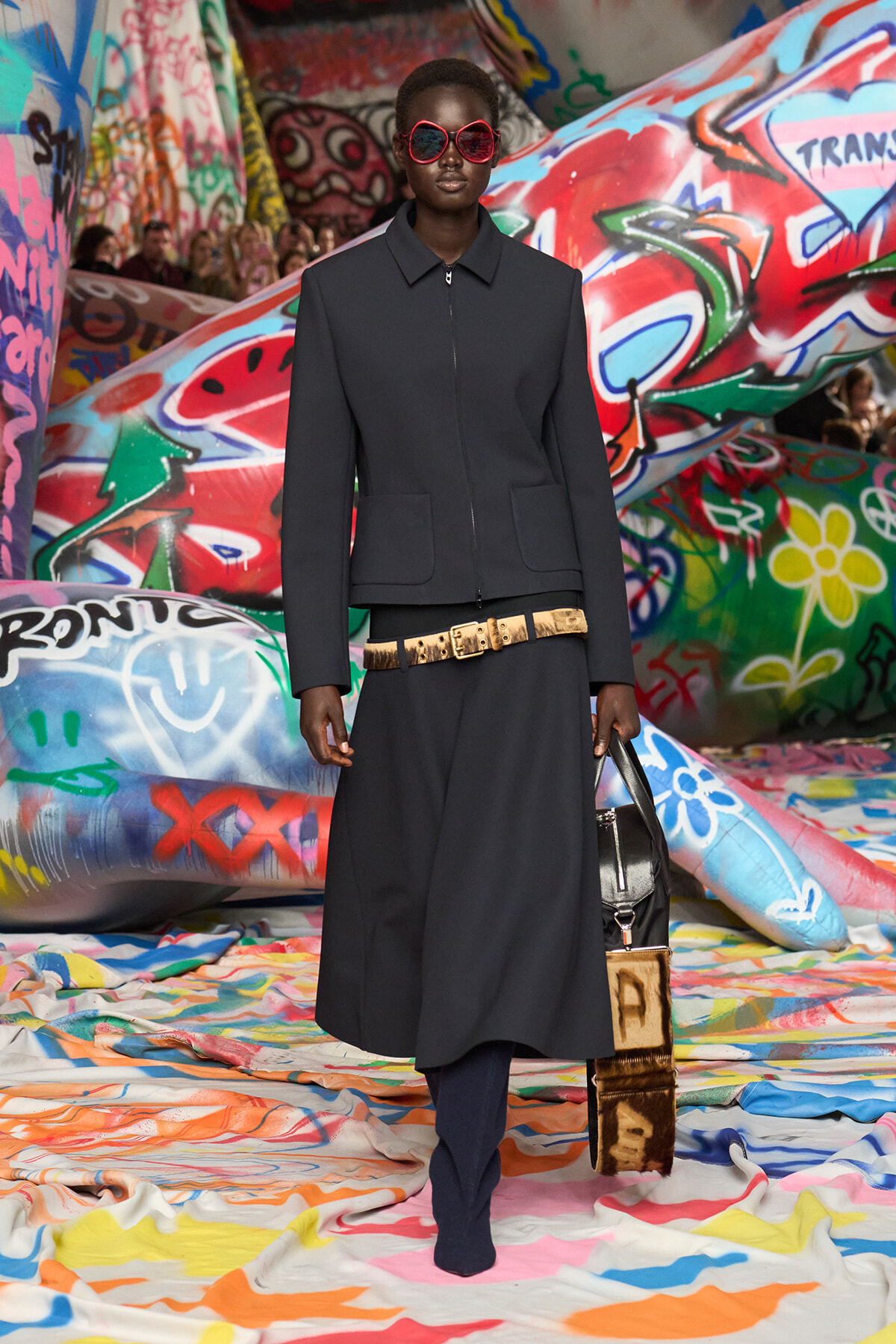 Model on a graffiti-covered runway wearing a dark zip-front jacket, red sunglasses, a wide gold belt, navy trousers, and a black bag.