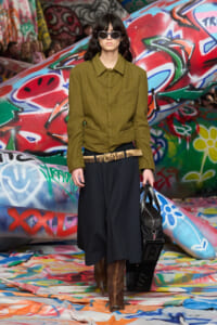 Model on a graffiti-covered runway wearing an olive-green jacket, black wide-leg skirt, brown knee-high boots, and sunglasses, carrying a black bag.