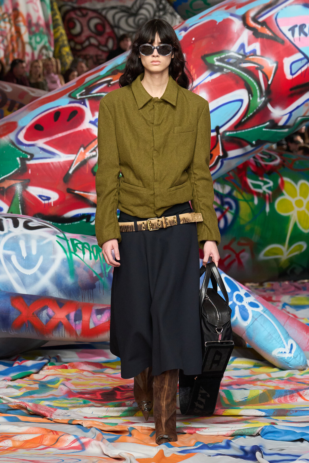 Model on a graffiti-covered runway wearing an olive-green jacket, black wide-leg skirt, brown knee-high boots, and sunglasses, carrying a black bag.