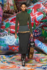 Runway model walking a graffiti-covered set in a green ribbed turtleneck, blue-gray waist cincher, charcoal midi skirt, tall black boots, and a black bag with brown accents.