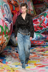 Model walking through a graffiti-covered tunnel in a black leather jacket, gray top, baggy jeans, and a metallic belt.