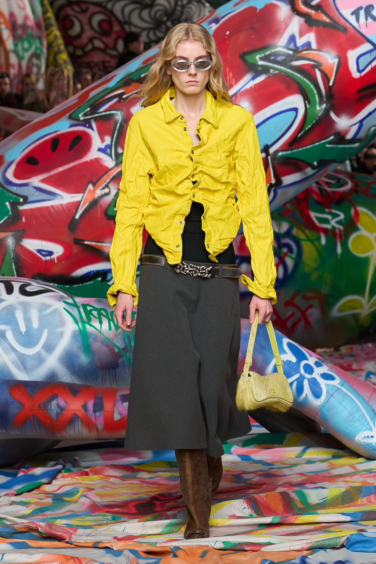 Model on a graffiti-covered runway wearing a bright yellow button-down shirt, dark wide-leg skirt, brown knee-high boots, belt, sunglasses, and a yellow handbag.