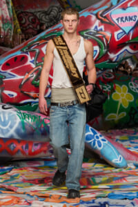 Male model walking a graffiti-covered runway in a white tank top, faded jeans, a wide belt, and a brown sash across his chest, carrying a black bag.