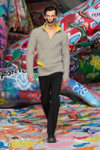 Male model striding down a graffiti-covered runway in a gray textured pullover and black pants with a yellow accent in the sweater edges.