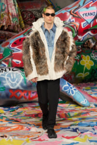Model walking runway in a cream parka with fur panels, denim shirt, and sunglasses against a graffiti-covered backdrop.