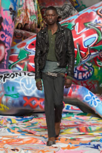 Male model walking a fashion show wearing a black leather jacket over a green knit top and gray trousers, with a braided belt, on a graffiti-covered runway backdrop.