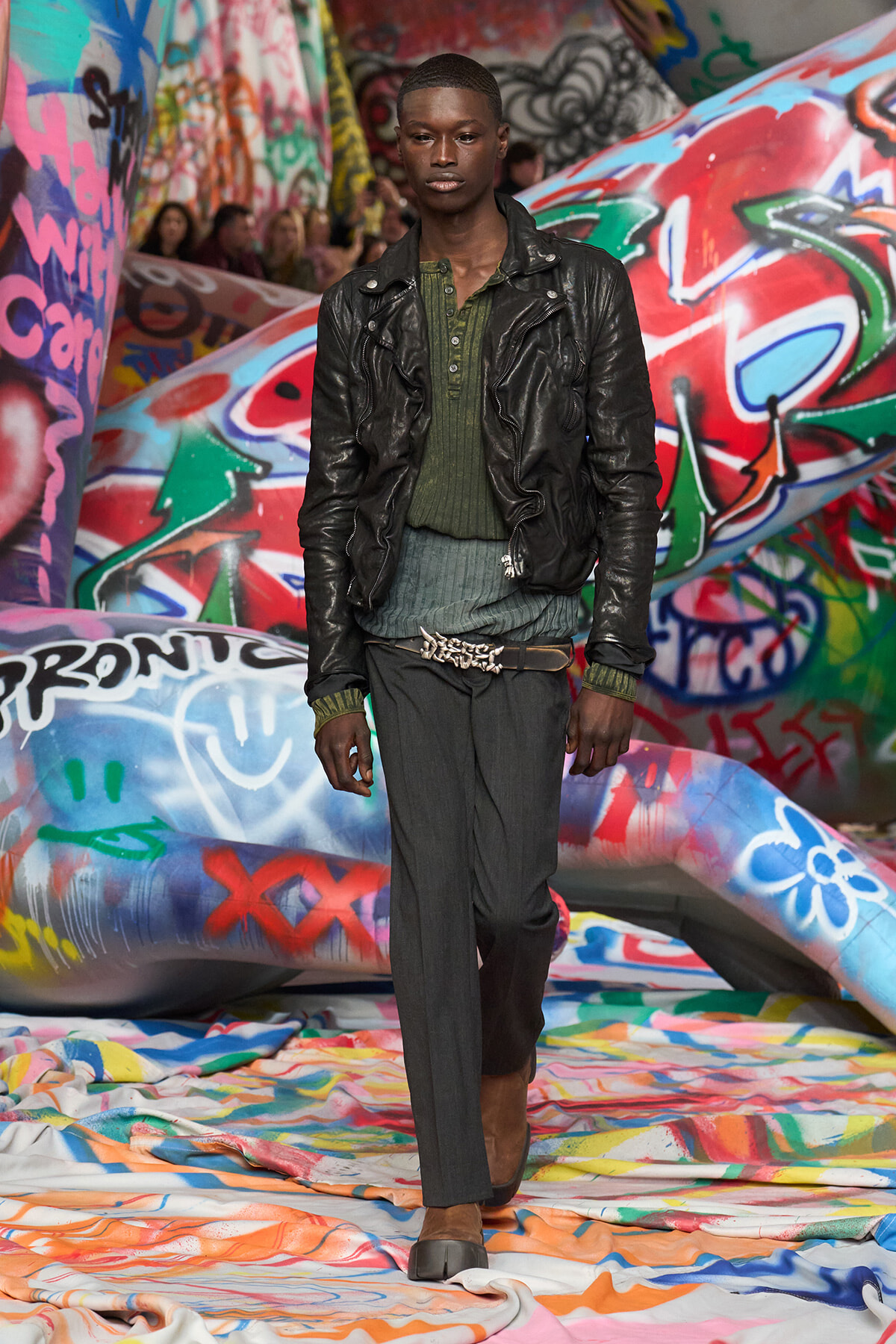 Male model walking a fashion show wearing a black leather jacket over a green knit top and gray trousers, with a braided belt, on a graffiti-covered runway backdrop.