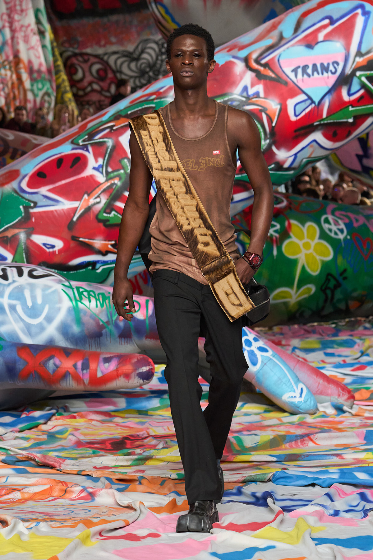 Male runway model walking on a graffiti-covered catwalk, wearing a brown sleeveless shirt, black pants, black shoes, and a wide tan sash across his torso.