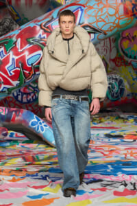Young male model in an oversized beige puffer jacket and baggy jeans, standing in a graffiti-covered space with colorful walls and ground spray paint.